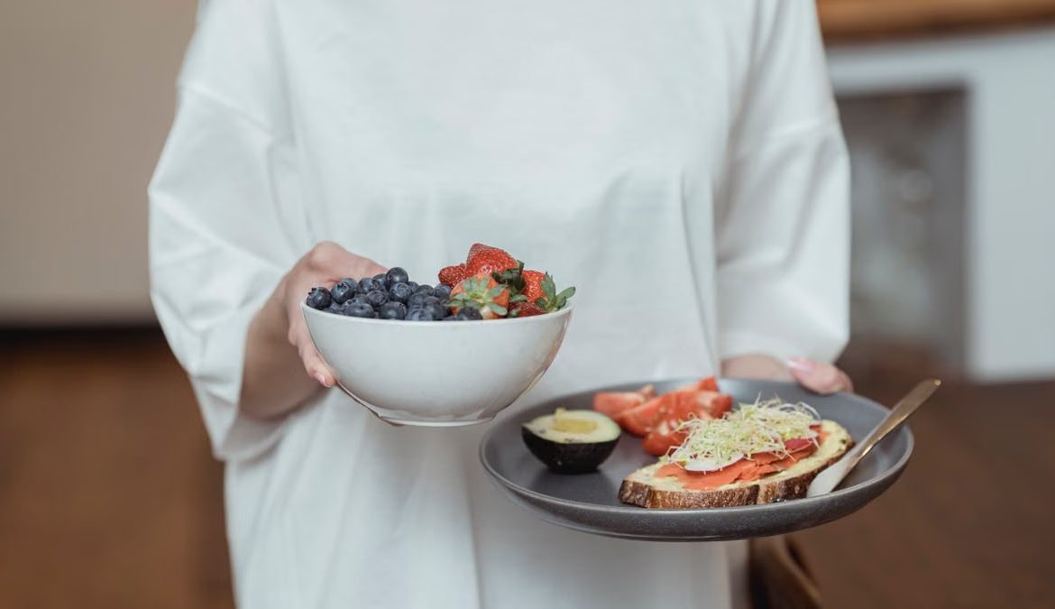 Person holding a bowl of fresh berries and a plate with avocado toast, displaying a healthy breakfast spread.